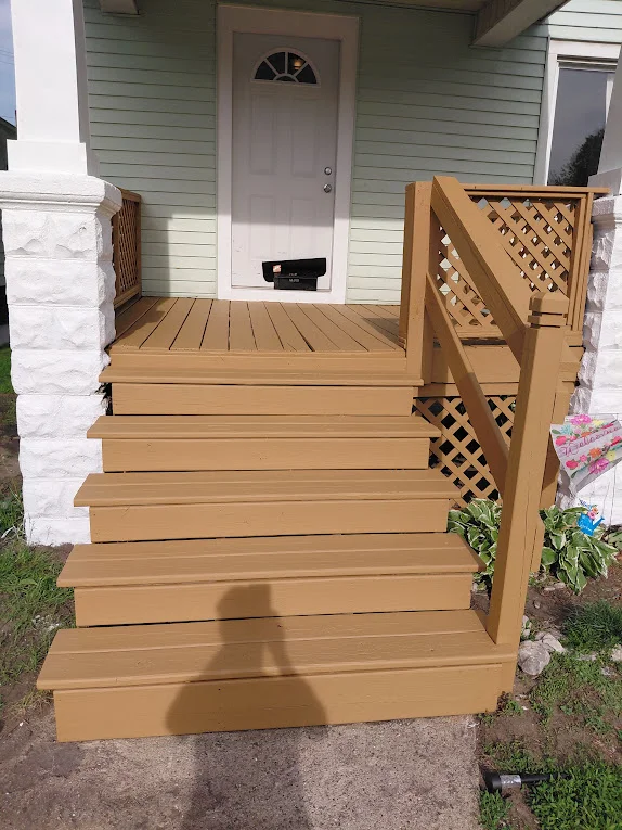 Home entrance with light green exterior, a wooden deck with stairs, and a decorative lattice railing leading to a white door