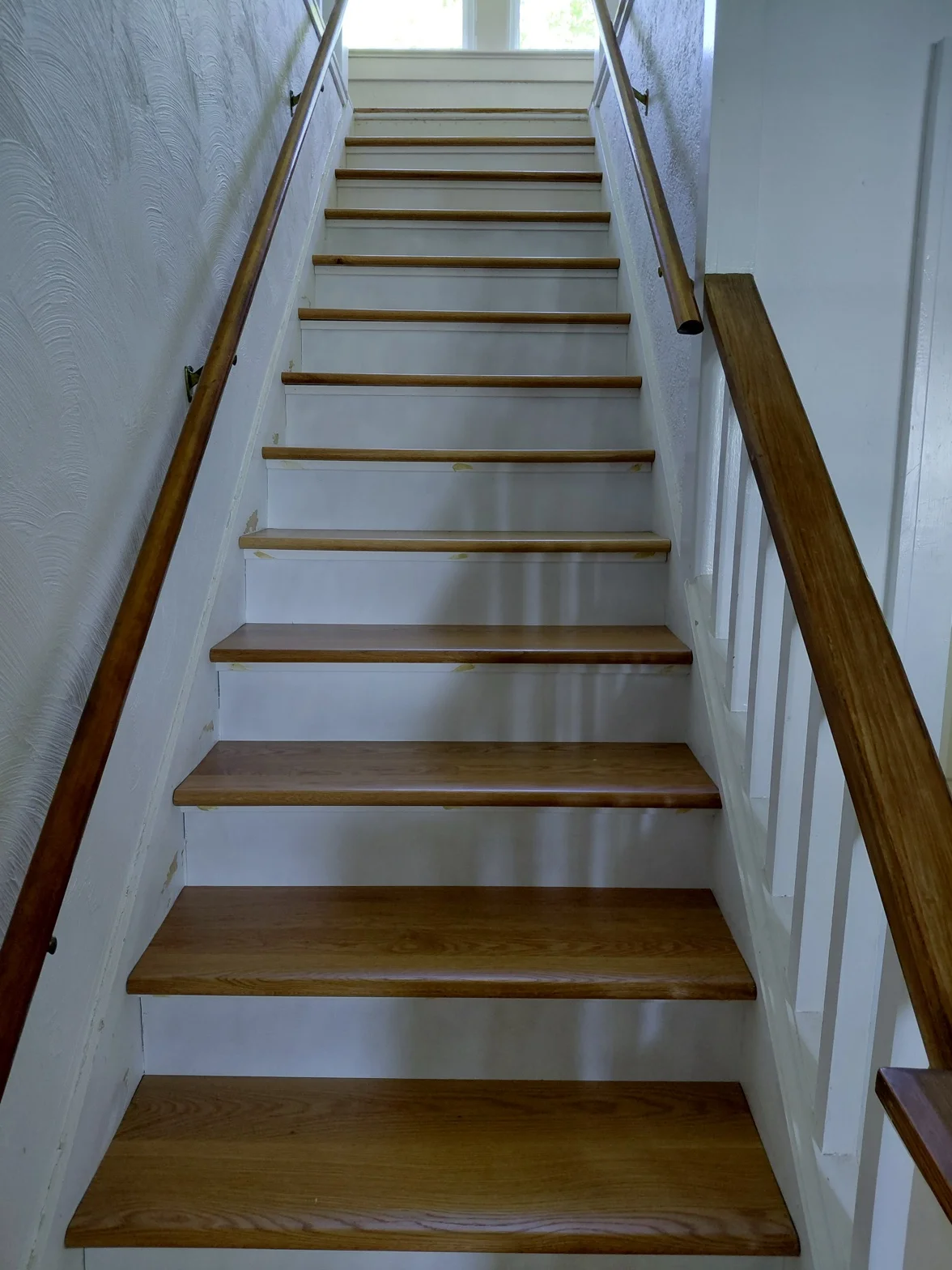 Well-lit staircase with white walls and wooden steps, viewed looking upward toward natural light